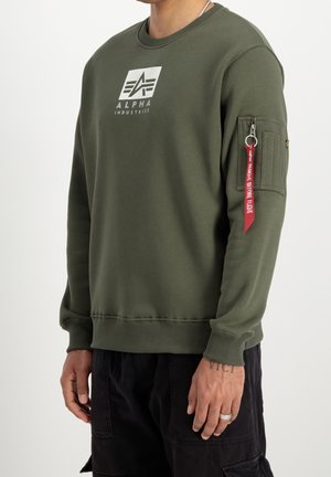 Mężczyzna noszący oliwkową bluzę Alpha Industries z logo na piersi oraz kieszenią na rękawie na zamek błyskawiczny z czerwoną zawieszką "Remove Before Flight".