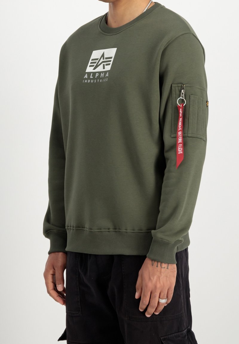 Homme portant un sweat-shirt vert olive Alpha Industries avec logo sur la poitrine et une poche zippée sur la manche avec une étiquette rouge « Remove Before Flight ».