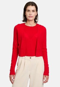 Gilet rouge court avec des manches longues, texture côtelée, design ouvert à l'avant, superposé sur un haut rouge ajusté, associé à un pantalon de couleur crème.