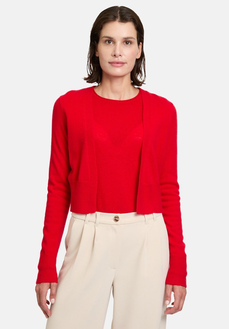 Gilet rouge court avec des manches longues, texture côtelée, design ouvert à l'avant, superposé sur un haut rouge ajusté, associé à un pantalon de couleur crème.