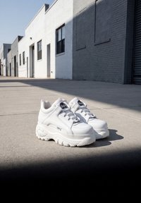 Paire de baskets blanches épaisses posées sur un trottoir en béton ensoleillé, avec des bâtiments en briques blanches et grises en arrière-plan.