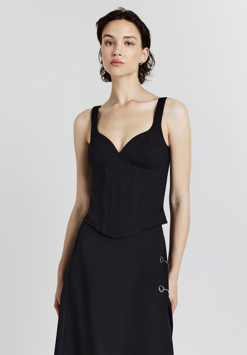 Schwarzes Korsett-Top mit breiten Trägern, Herzausschnitt, strukturierten Nähten und glatter Textur. Kombiniert mit einem schwarzen Rock mit runden Akzenten.