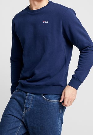 Mann trägt ein marineblaues FILA-Sweatshirt und blaue Jeans, mit einer Hand nahe der vorderen Hosentasche.
