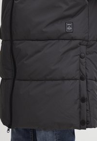 Veste matelassée noire avec fermeture éclair, matelassage visible et cinq boutons noirs à l'avant. Comprend un petit patch de marque sur la poitrine.