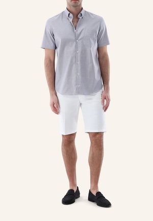 Mann trägt hellgraues kurzärmeliges Hemd mit Knöpfen, weiße kniehohe Shorts und schwarze Loafer, steht vor einfachem Hintergrund.