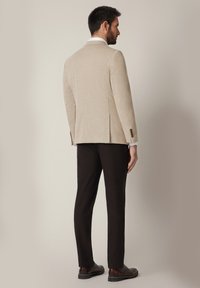 Mężczyzna stojący bokiem, ubrany w beżową marynarkę, białą koszulę, czarne spodnie i brązowe buty eleganckie, na jednolitym tle.