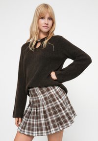 Brauner gestrickter Pullover mit einem Schlüssel-Ausschnitt, kombiniert mit einem gefalteten Karorock in Weiß-, Braun- und Schwarztönen. Sichtbares Detail des strukturierten Stoffes.