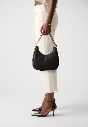 Borsa a tracolla nera in pelle con forma curva, superficie testurizzata, tracolla in catena dorata e base piatta. Abbinata a pantaloni beige a vita alta e tacchi neri.