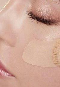 Beige flytende foundation påføres med en pensel på glatt hud; fremhever hudens tekstur og sminkens blanding. Nærbilde av ansiktet.