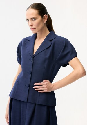 Kvinde med bagstrøget hår iført en kortærmet marineblå blazer med tre knapper, poserer med den ene hånd på hoften.