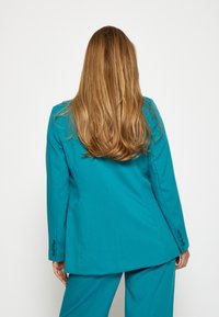 Tirk�īza žakete ar teksturētu audumu, ar vienu aizmugurējo venti, un trijām pogām pie piedurkņu galiem, uzvilkta kopā ar atbilstošām biksēm, skatoties no aizmugures.