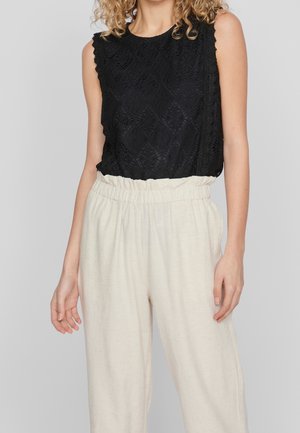 Femme portant un haut noir en dentelle sans manches, rentré dans un pantalon ample taille haute de couleur blanc cassé avec une ceinture élastique, se tenant devant un fond gris.