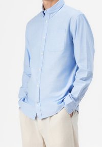 Camisa de botones de manga larga de color azul claro en una tela suave. Presenta un bolsillo en el pecho, dobladillo redondeado y puños con botones. Se lleva con pantalones beige.