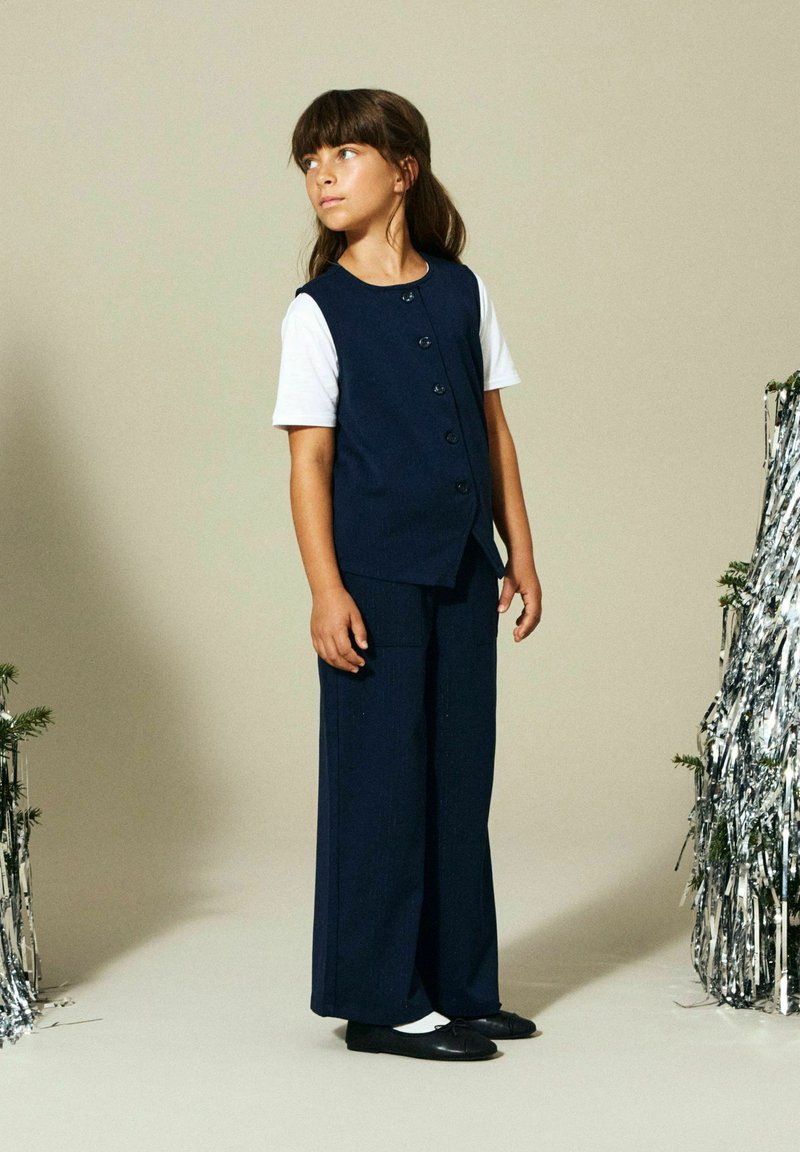 Temno modna dvodelna obleka z brezrokavnim telovnikom z gumbi in širokimi hlačnicami. Gladek material s subtilno teksturo. Črne balerinke na nogah.