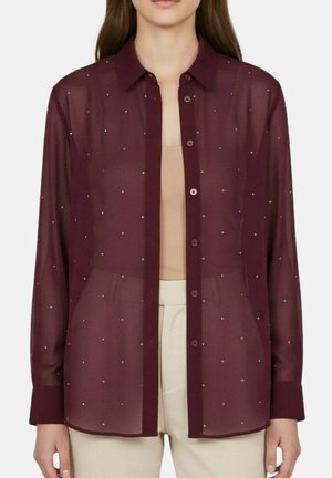 Femme portant un chemisier bordeaux transparent à petits pois blancs sur un top beige et un pantalon blanc.