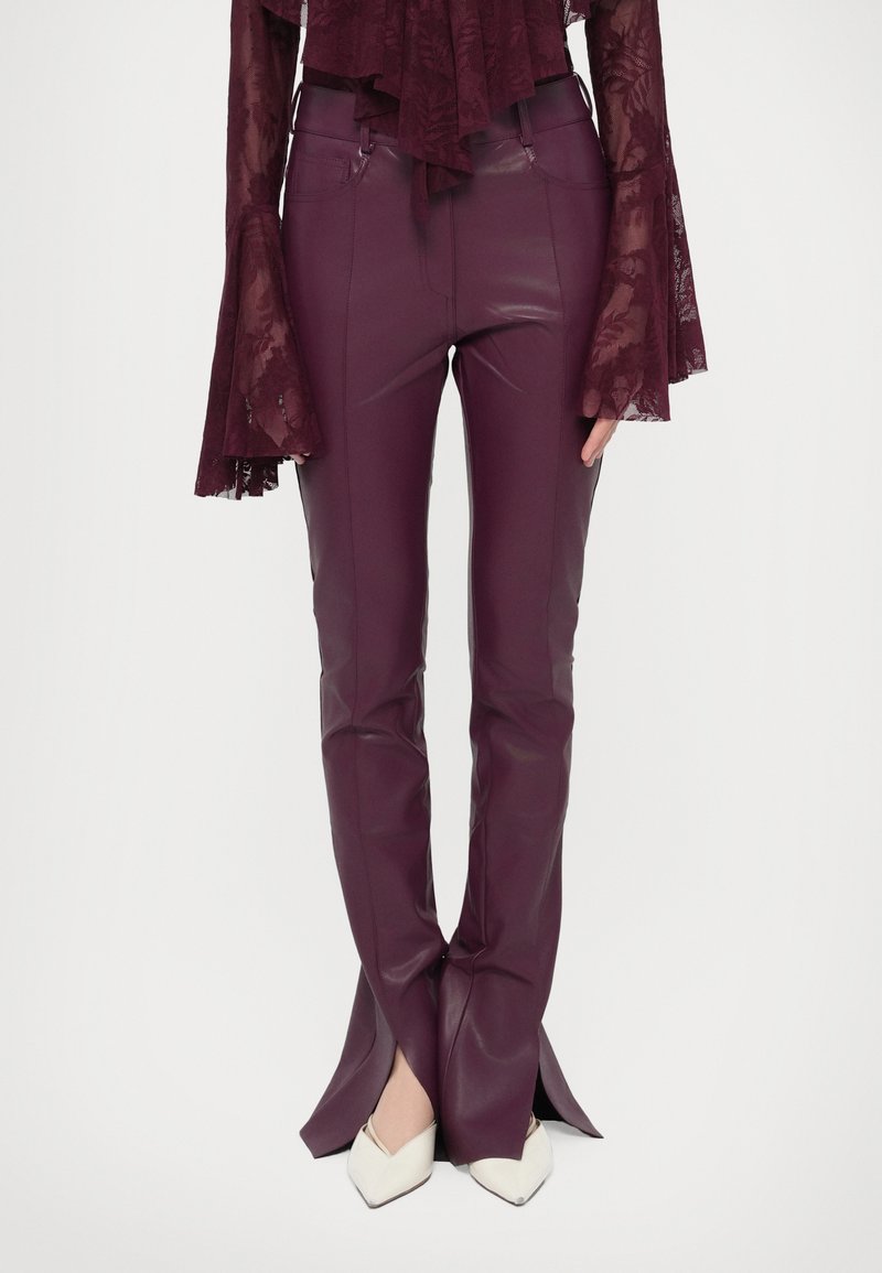 Bordó műbőr nadrág, testhez simuló kialakítással, az alsó részen oldalsó hasítékokkal és sima, fényes textúrával.