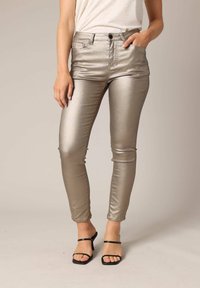 Metaalachtige zilveren skinny jeans met een gladde, glanzende textuur, voorzien van een sluiting met een voorknoop en zakken, gecombineerd met zwarte bandjesandalen.