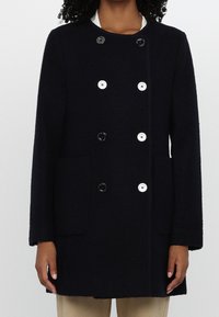 Femme portant un manteau double boutonnage en laine noire avec des boutons alternant noir et blanc ainsi que des poches plaquées à l'avant, pantalon beige visible.