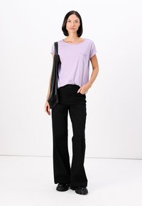 Lila korte mouwen t-shirt met een borstzak, gecombineerd met zwarte wijde broek en chunky zwarte schoenen. Model houdt een zwarte tote tas vast.