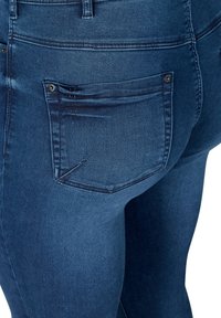 Jean en denim bleu foncé avec une texture lisse, taille haute, et une poche arrière décorée avec des détails de couture. Vue rapprochée de l'arrière.