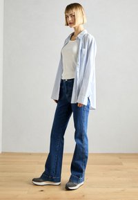 Camisa de botones a rayas azul claro sobre una camiseta blanca sin mangas, combinada con jeans acampanados azul oscuro y zapatillas grises. Un atuendo simple y casual.