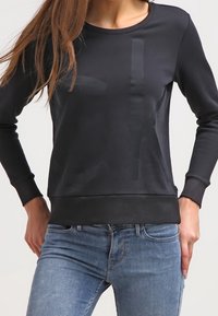 Svart sweatshirt med rundad halsringning, långärmade, ribbade muddar och en diskret logodesign på framsidan. Tyget verkar slätt och tight.