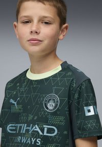 Zöld, rövid ujjú futballmez, háromszögmintákkal, világos zöld gallérral, valamint a Manchester City logójával és brandinggel az elülső oldalon.