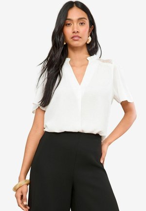 White short-sleeve blouse with a v-neckline, featuring textured shoulder accents and a relaxed fit, paired with black trousers.