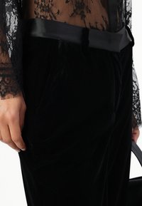 Sorte fløyelsbukser med en glatt tekstur og satengliv, paret med en gjennomsiktig svart spissbluse med blomsterdekorasjoner.