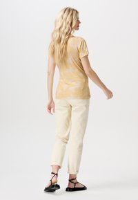 Geel gemusterd t-shirt met korte mouwen, gecombineerd met licht roomkleurige broek. Zwarte platte sandalen met kruislings gespen. Neutrale achtergrond.