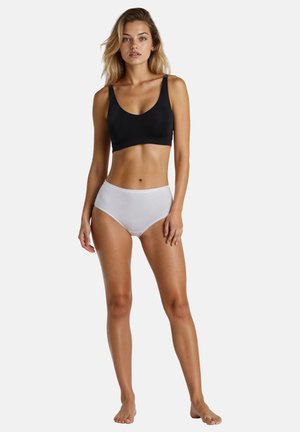 Femme debout pieds nus portant un soutien-gorge de sport noir et un slip taille haute blanc sur un fond clair uni.