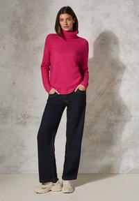 Fuchsia-Strickpullover mit hohem Kragen, kombiniert mit dunkelblauen weiten Jeans und beigen Sneakern. Weicher Stoff und figurbetonte Silhouette.