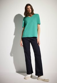 Grön kortärmad t-shirt med V-ringning, kombinerad med mörkblå raka jeans och vita sneakers. Slät tygtextur.