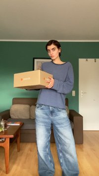 Caixa de cartão com o logotipo da Zalando, segurada por uma pessoa vestindo uma camisa de manga longa com riscas azuis e brancas e calças de algodão largas de cor azul clara.