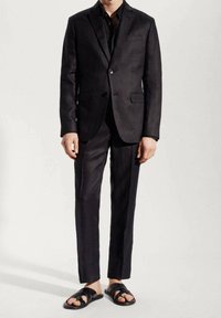 Man wearing a black suit with a black shirt and black open-toe sandals, standing against a plain light background.