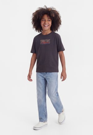 Uśmiechnięte dziecko z kręconymi włosami, ubrane w czarną koszulkę z napisem „Sunny Days California”, jasnoniebieskie dżinsy oraz białe buty wsuwane typu slip-on, stojące na białym tle.