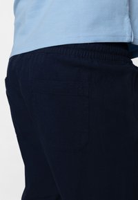 Pantalon bleu marine avec taille élastique, une poche arrière unique et une texture de tissu douce, partiellement visible sous un haut bleu clair.