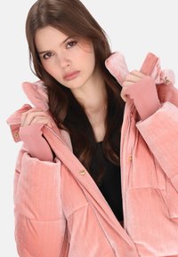 Różowa pikowana kurtka puchowa z wysokim kołnierzem, złotymi elementami i różowymi ściągaczami. Materiał ma welwetową fakturę z subtelnymi paskami.