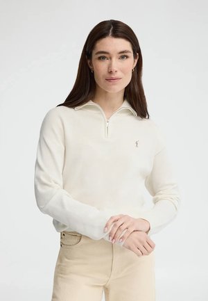 Kobieta o długich brązowych włosach, ubrana w kremowy sweter zapinany na zamek i beżowe spodnie, stojąca z lekko splecionymi dłońmi na białym tle.