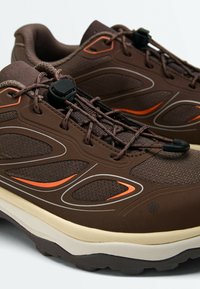 Paire de chaussures de sport marron avec des accents orange, tissu texturé et système de laçage rapide sur fond blanc.