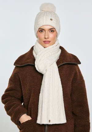 Naine, kes kannab pruuni fliisjopet, käed taskutes, kaela ümber on kerge kreemikaabli mustriga sall ning peas sobiv tutiga müts.