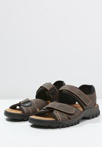 Braune Sandalen mit Klettverschlussriemen, ausgestattet mit einer gepolsterten Korkinnensohle und einer strukturierten Gummisohle in Schwarz. Kontrastnähte setzen Akzente.
