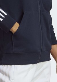 Person som bär en marinblå zip-up jacka med handen i framfickan, tillsammans med vita byxor med svaga ränder.