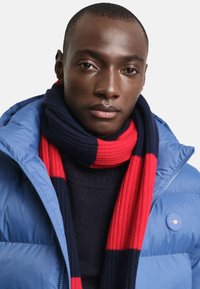 Modrá péřová bunda s kulatým logem, námořnický a červený pruhovaný žebrovaný šátek, pod ním strukturovaný pletený svetr tmavé barvy, hladká pleť.