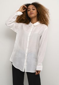 Donna con capelli ricci che indossa una camicia bianca a bottoni e pantaloni neri, in posa con una mano che tocca la testa su uno sfondo semplice.
