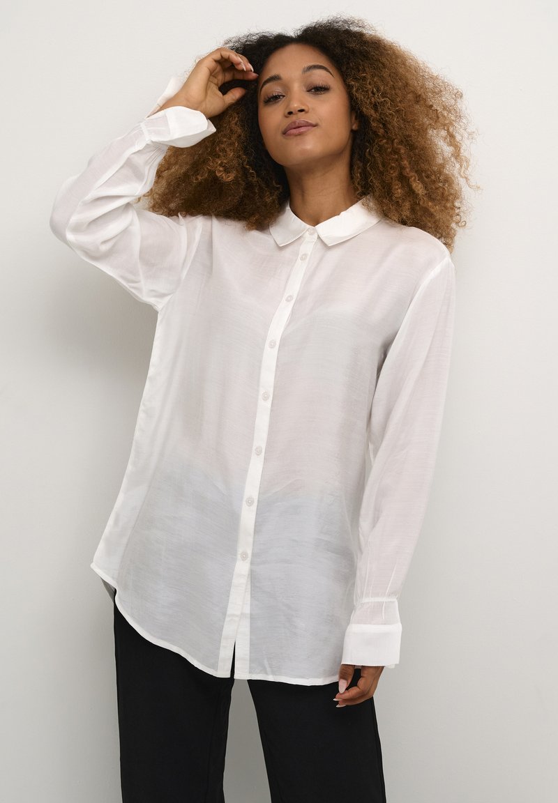Donna con capelli ricci che indossa una camicia bianca a bottoni e pantaloni neri, in posa con una mano che tocca la testa su uno sfondo semplice.