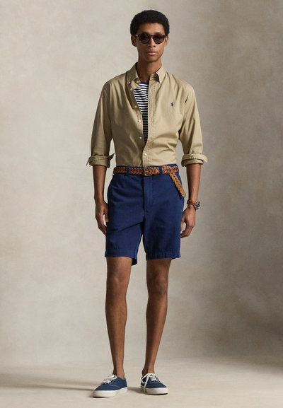Άνδρας που φοράει γυαλιά ηλίου, μπεζ πουκάμισο με κουμπιά πάνω από ριγέ μπλουζάκι, μπλε σορτς με πλεγμένη ζώνη, μπλε αθλητικά παπούτσια, στέκεται μπροστά σε απλό φόντο.