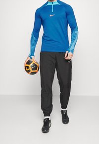 Sininen pitkähihaisen urheilu- paita, jossa on puolivetoketju ja geometrisesti kuvioidut hihat, yhdistettynä mustiin housuihin. Mustat jalkapallositeet ja pallo.