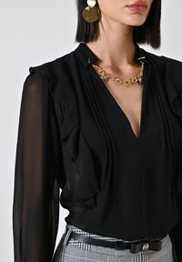 Blusa nera a maniche lunghe con dettagli arricciati, scollatura a V e collana con catena dorata. Abbinata a pantaloni a scacchi e grandi orecchini dorati.