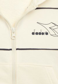 Felpa con cappuccio color crema con zip, dettagli neri, con logo Diadora e interno testurizzato. Include una tasca frontale e orlo a coste.